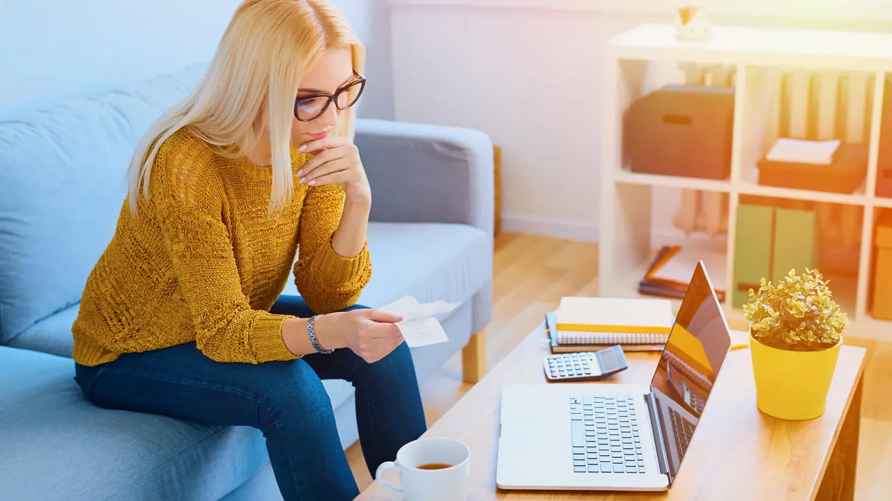 young woman counting expenses at home with bills and laptop sitting on sofa.