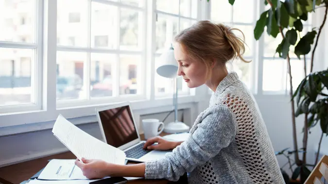 Woman looking at document and laptop next to window filtering in natural light