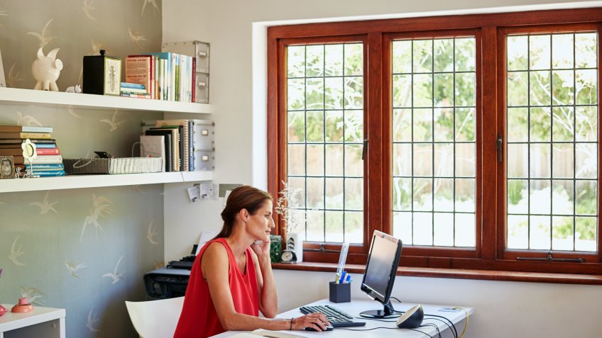 Mid adult woman using computer and mobile phone at desk in house.