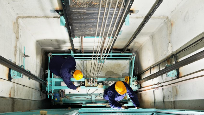 two male technician machinist worker at work adjusting elevator mechanism of lift with spanner.