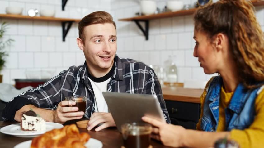 Smiling handsome young man talking to friend and drinking coffee from mug at home, they using tablet to watch video.