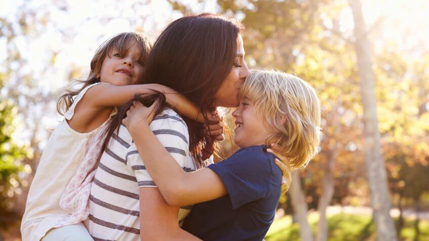 Mother Carrying Son And Daughter As They Play In Park.