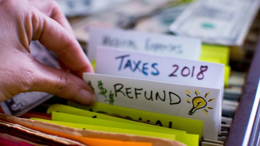 Hand pulling out file in filing cabinet with tax forms and papers with words Refund and Taxes 2018 handwritten on file folder conceptual dollar signs images and light bulb for ideas on tax refund conceptual financial planning photography.
