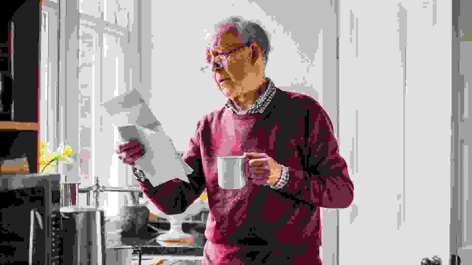 Senior man is standing in the kitchen of his home with bills in one hand and a cup of tea in the other.