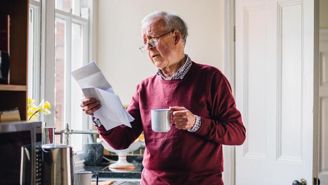 Senior man is standing in the kitchen of his home with bills in one hand and a cup of tea in the other.
