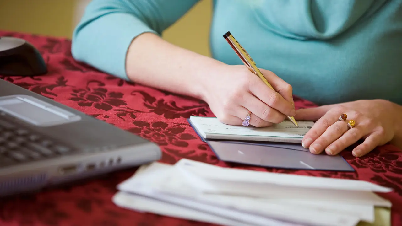 Woman sitting at her table next to a laptop going over the budget and writing a check in her checkbook