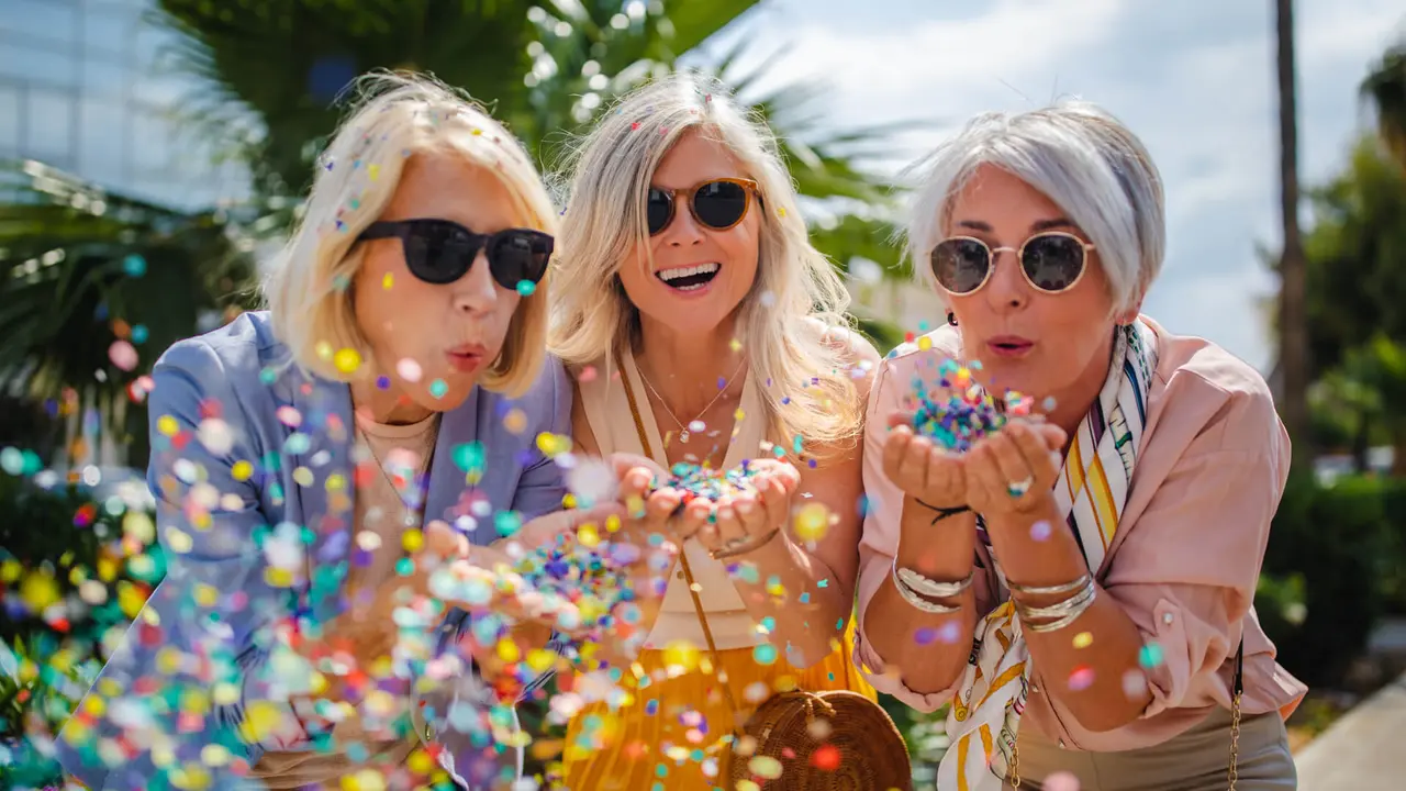Fashionable mature friends having fun and celebrating by blowing colorful confetti in city street.