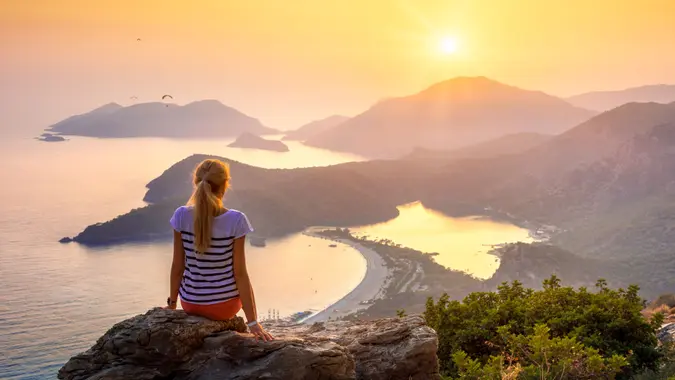 Young woman sitting on the top of rock and looking at the seashore and mountains at colorful sunset in summer.