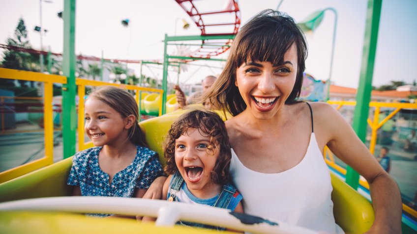 Excited little boy and girl having fun with mother riding amusement park roller coaster.