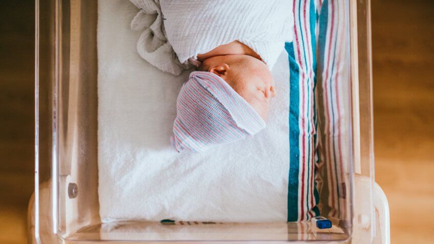 A baby just born at the hospital rests in a hospital bassinet crib, wrapped in a swaddle and wearing a beanie hat.
