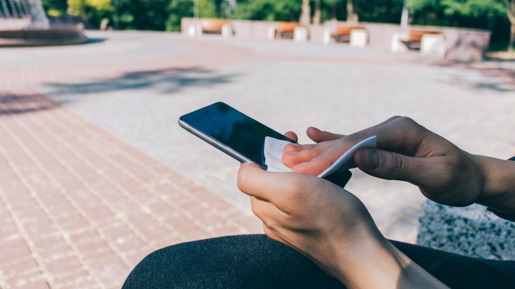 Woman cleaning display of smart phone with antibacterial white tissue, while sitting in the public park.