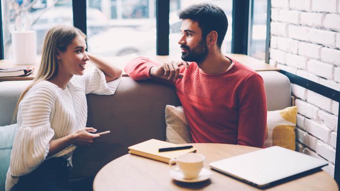 Young woman and man talking during first date in cozy coffee shop enjoying free time together,hipster girl checking notification on mobile while spending time with boyfriend having lovely conversation - Image.