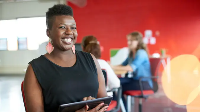 Casual portrait of an african american business woman using technology in a bright and sunny startup with the team in the background.