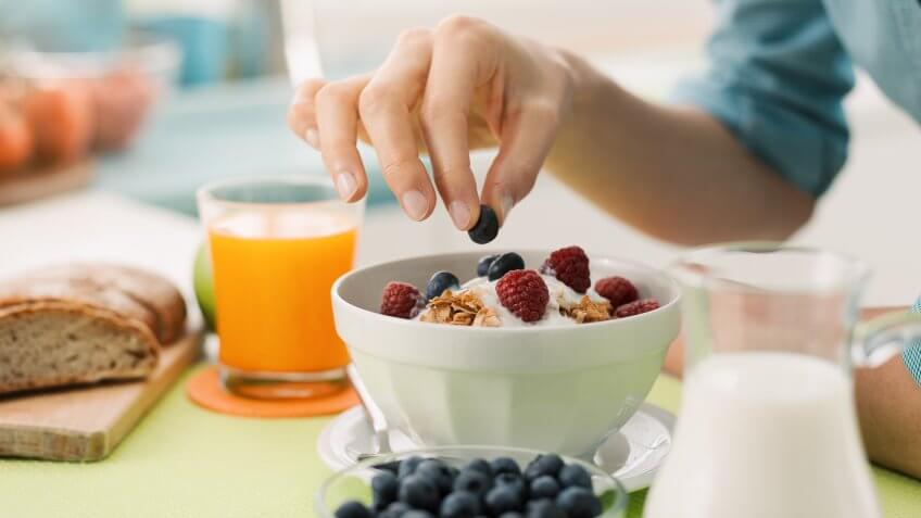 Woman having an healthy delicious breakfast at home with yogurt, cereals and fresh fruit, she is picking a blueberry.