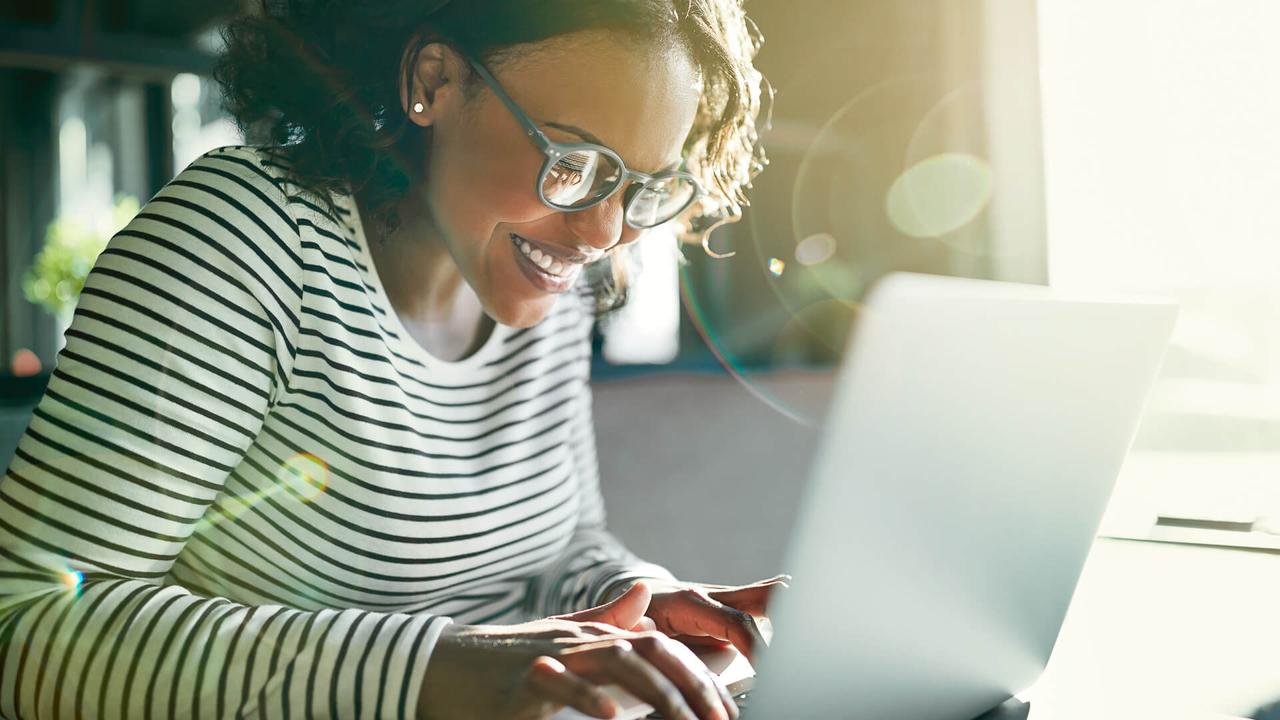 Young African woman wearing glasses smiling while sitting alone at a table working online with a laptop.