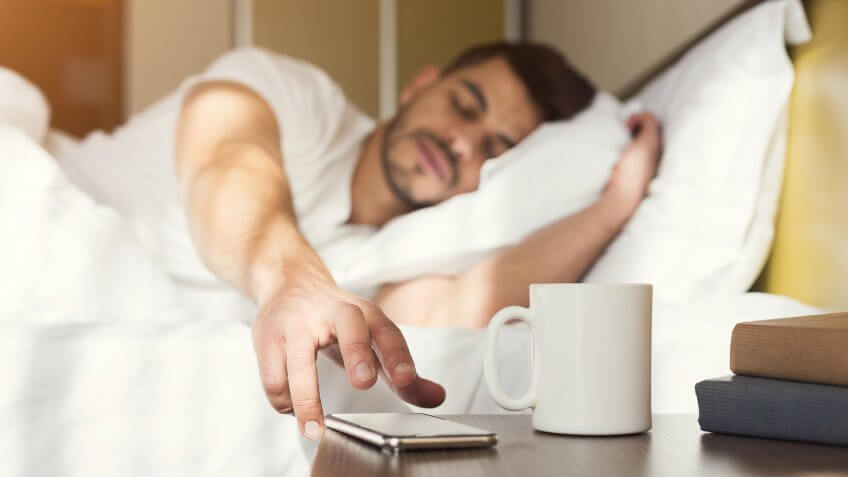 Sleepy guy waking up early after hearing alarm clock signal on smartphone on monday morning, reaching for ringing mobile phone with closed eyes, copy space.