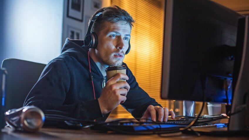 Portrait of focused beard programmer typing in digital device while tasting cup of coffee at night.