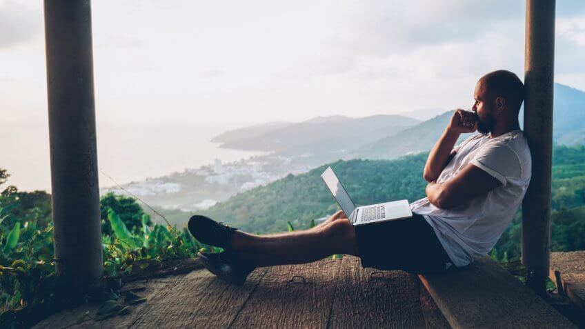 Thoughtful young man looking away on beautiful nature landscape while sitting with laptop computer outdoors.
