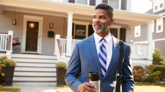 Businessman With Cup Of Coffee Leaving Suburban House For Work.