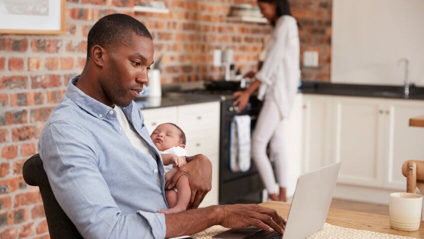 Father On Laptop Holds Newborn Son As Mother Makes Meal.