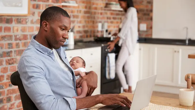 Father On Laptop Holds Newborn Son As Mother Makes Meal.