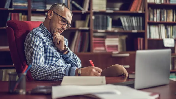 Mid adult manager sitting at home office and writing something in a notebook.