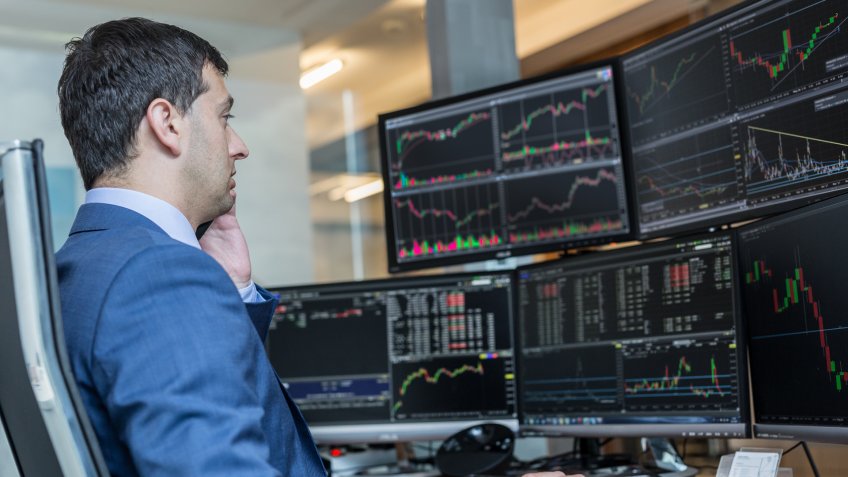 Male stock broker trading online watching charts and data analyses on multiple computer screens.