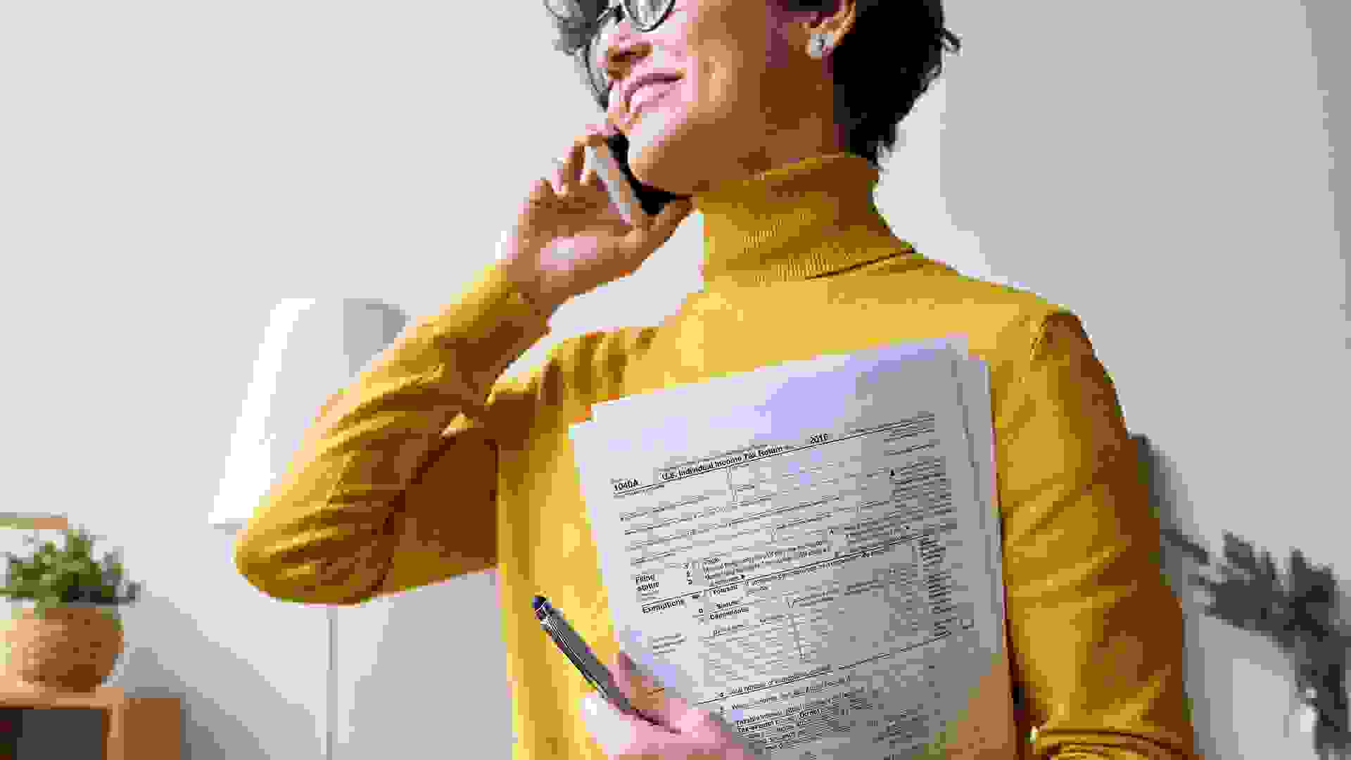 Cheerful woman in yellow sweater calling on phone and holding income tax return papers.