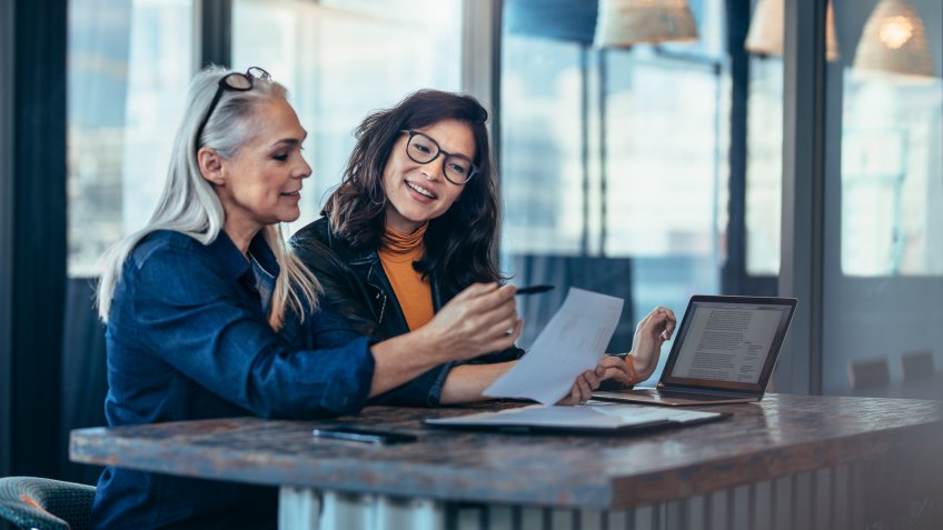 Two women analyzing documents while sitting on a table in office.