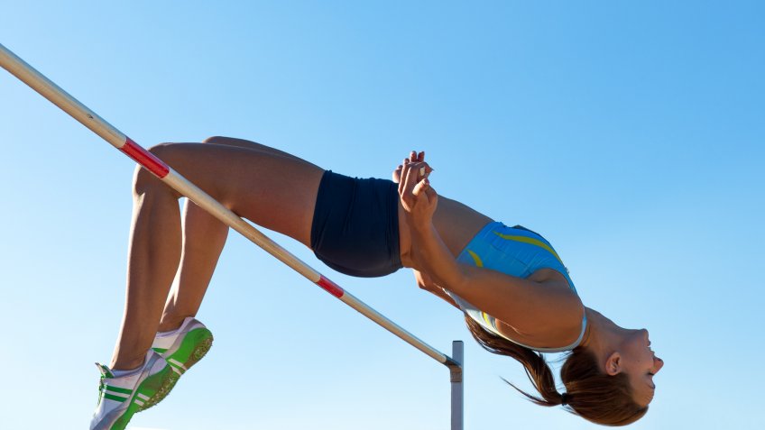 Side view of young female athlete jumping over the lath, against the sunlight.