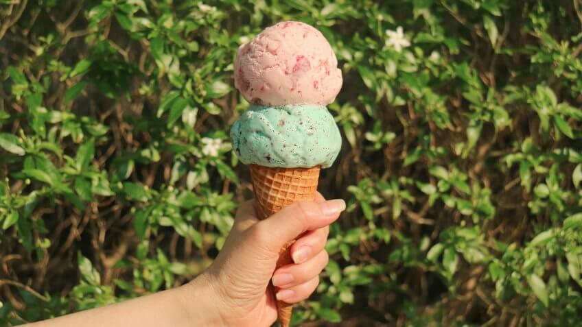 Hand holding an Ice cream cone with two scoops in the sunlight against blurry flowering tree.
