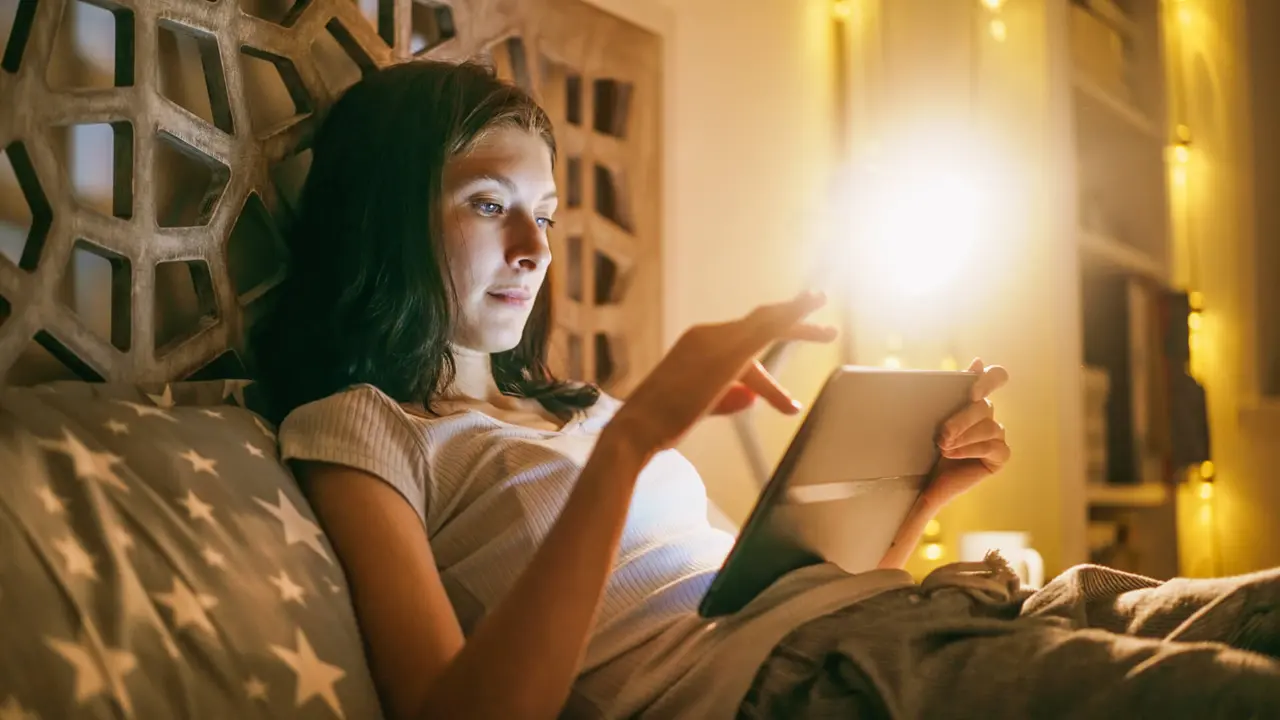 Young concentrated woman using tablet and watching online news lying in bed at home at night.
