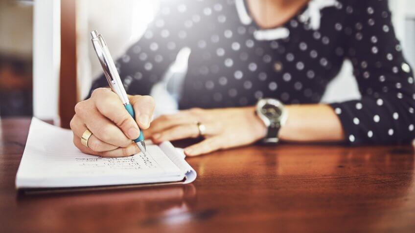 Shot of a unrecognizable woman writing in a book with a pen on a dinner table at home.