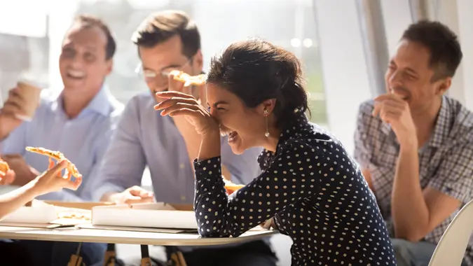 Indian woman laughing at funny joke eating pizza with diverse coworkers in office, friendly work team enjoying positive emotions and lunch together, happy colleagues staff group having fun at break.