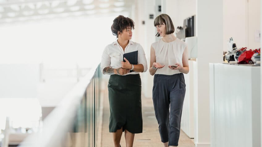 Two female professionals having a relaxed meeting while strolling along a raised walkway in an office auditorium.