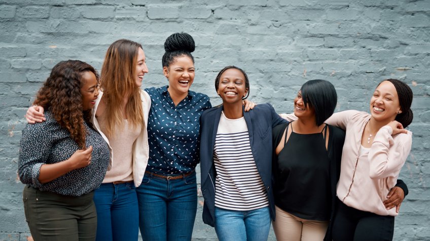 Portrait of a diverse group of young women standing together against a gray wall outside.