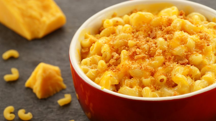 "Homestyle baked macroni and cheese in a red bowl with shredded cheese, bread crumbs, and cheese and pasta accents.