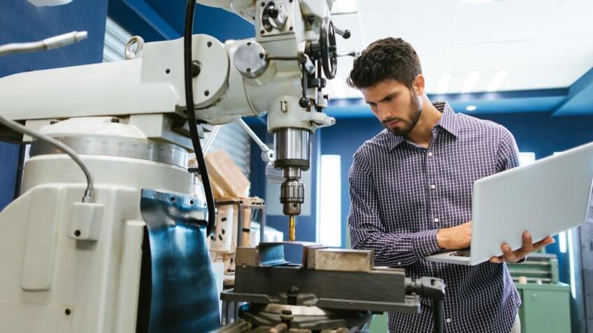 A young latin engineer calibrating a large drill with his laptop in his shop.