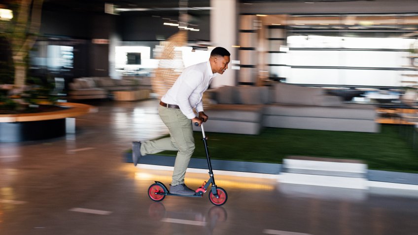 Young African businessman riding scooter inside shared office space.