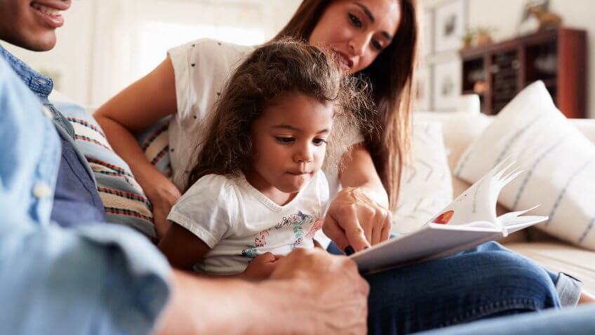 Hispanic couple and their young daughter sitting on the sofa reading a book together at home.