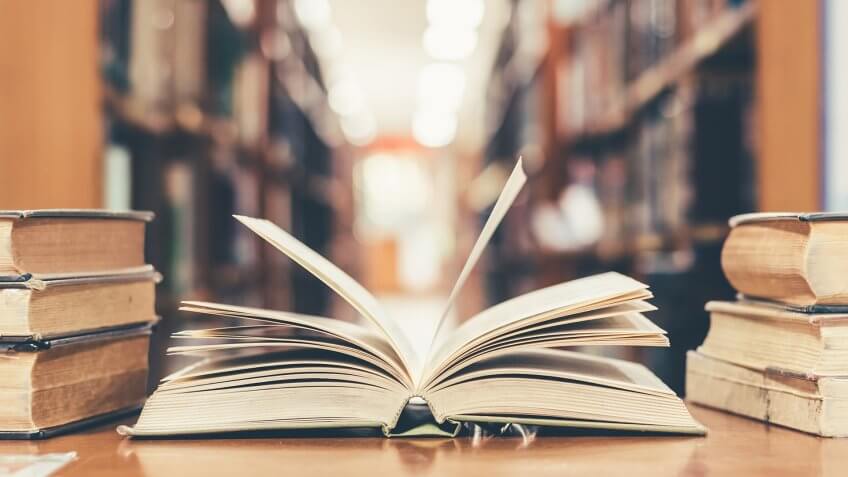 Education learning concept with opening book or textbook in old library, stack piles of literature text academic archive on reading desk and aisle of bookshelves in school study class room background - Image.