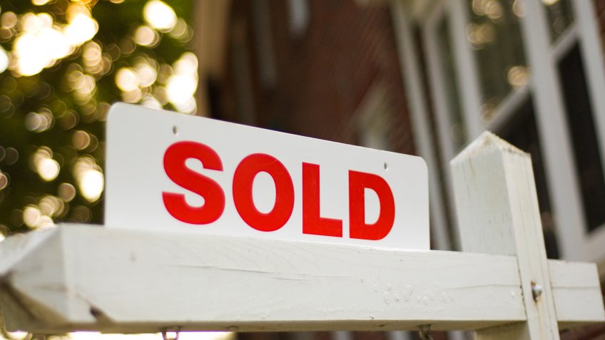 Real Estate "sold" sign with red brick building and trees blurry in the background.