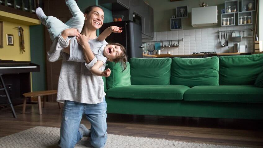 Excited mother holding lifting kid daughter flying laughing on weekend, young family single mom or babysitter nanny playing with active cute girl at home, happy mommy and child having fun together.