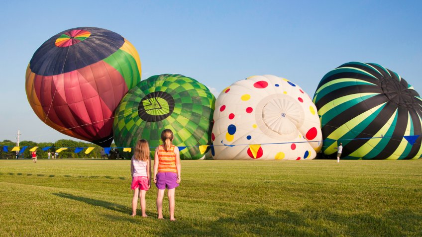 Four hot air balloons rise up into the blue sky at a festival in rural Kansas while two young girls watch from the grass - Image.