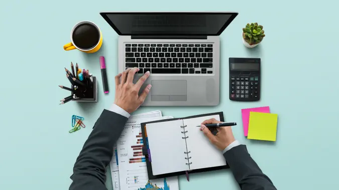 Office workplace with laptop, notebook, hand, office supplies, on turquoise background.