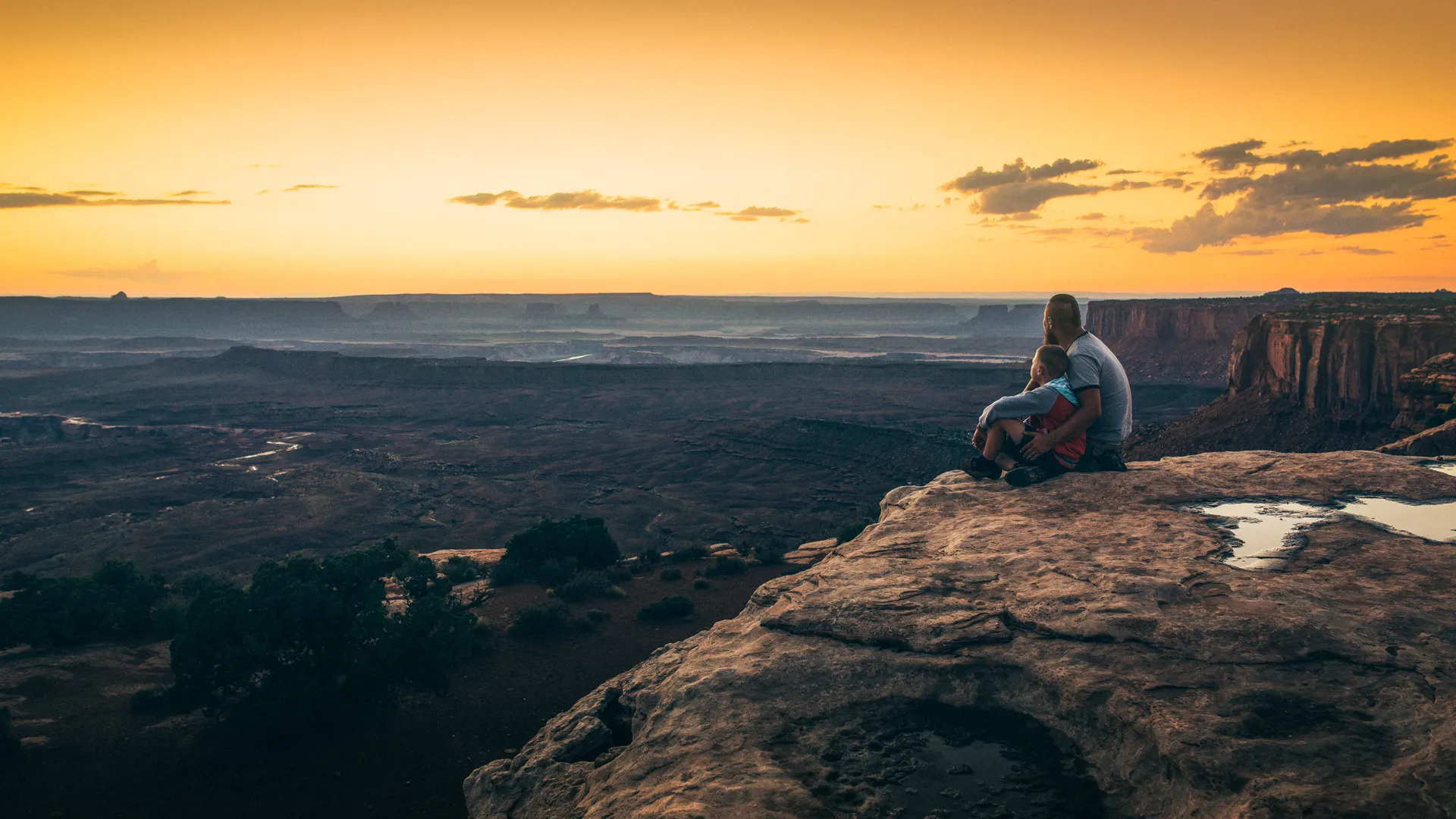 Father and son sitting on the edge of cliff overlooking vast landscape at Grand View Point of Canyonlands National Park during sunset.