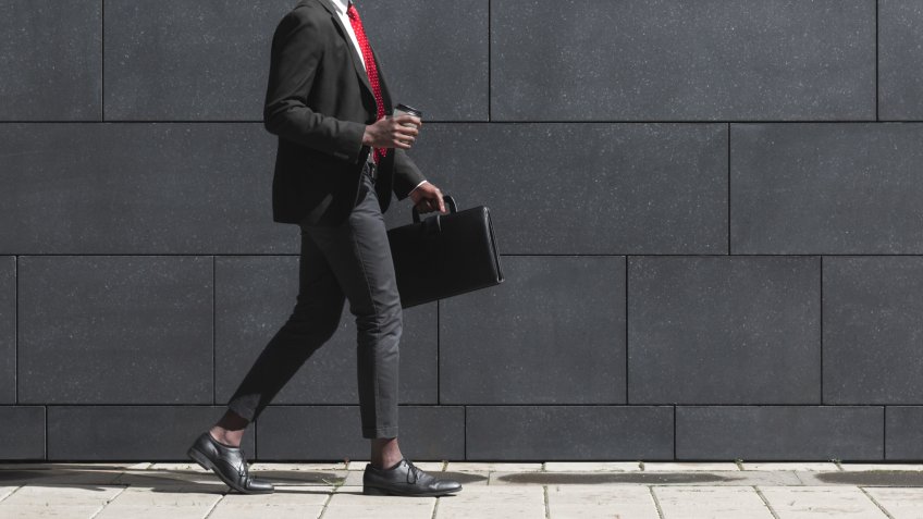 Full-length portrait of handsome African American entrepreneur walking on pavement with gray block wall in background, dressed in black suit, holding leather bag and coffee cup during break in work.