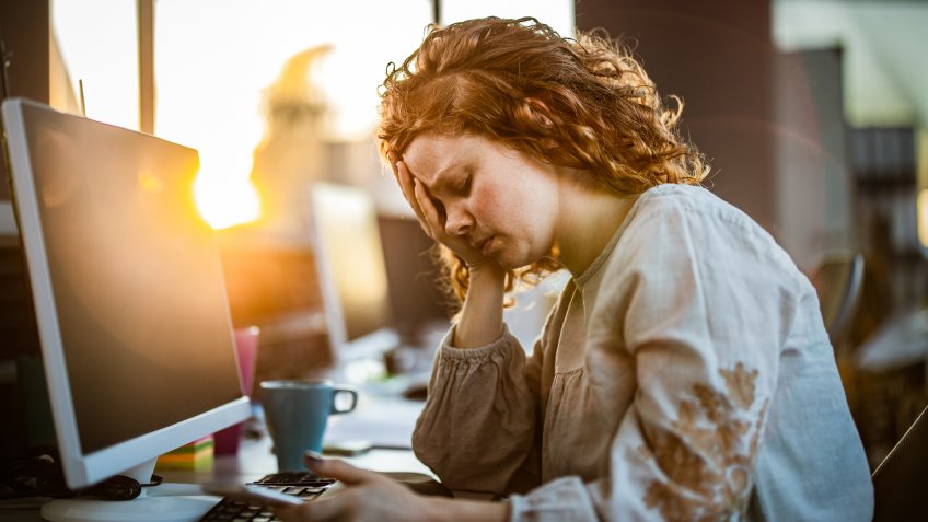 Exhausted redhead programmer holding her head in pain while working in the office.