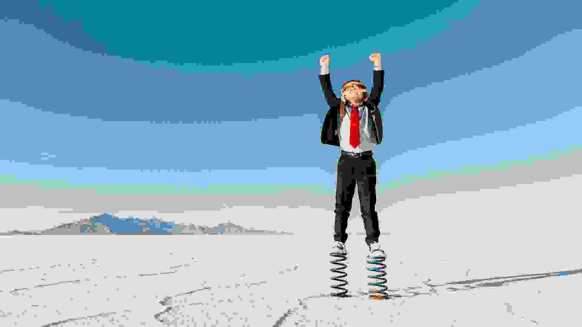 A young business boy dressed in business suit, flight cap and goggles stands on springs in the Utah desert.