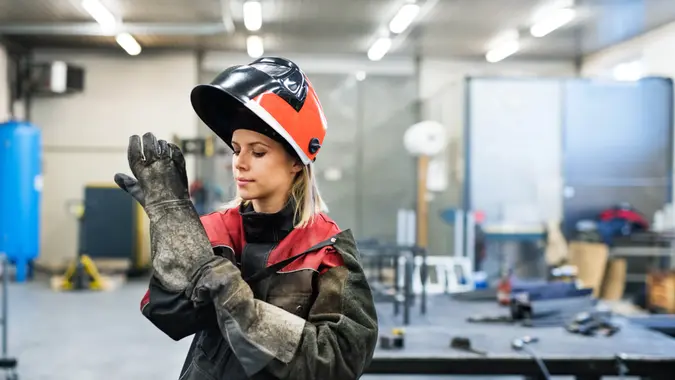 Industrial young woman worker at the welding factory, putting on protective gloves.